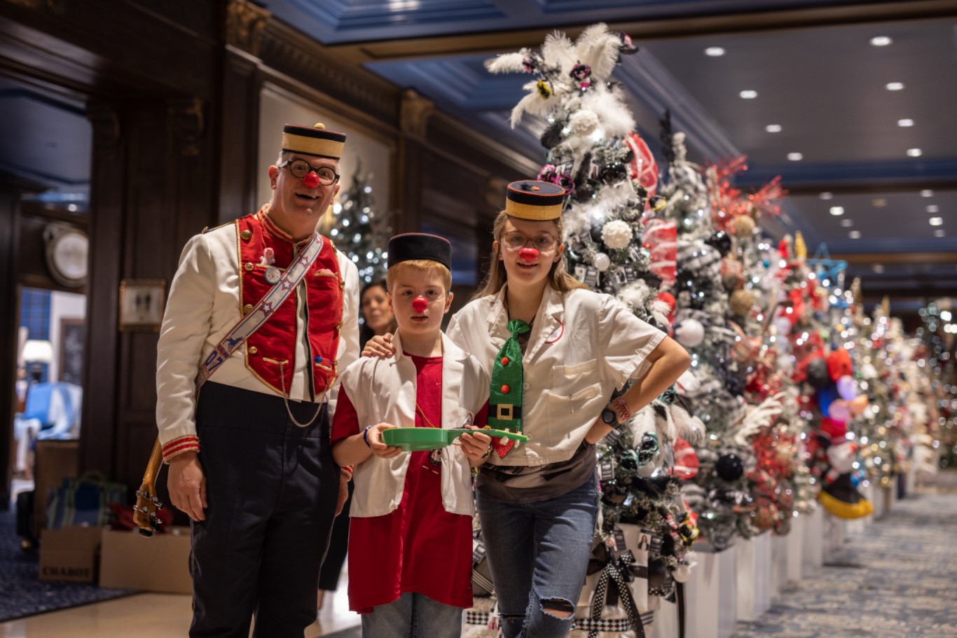 Man and two children with clown noses in festive attire in front of a line of beautifully decorated holiday trees of hope