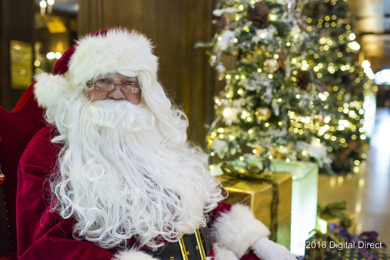 Santa Claus sitting in a red chair in front of presents and a Christmas tree